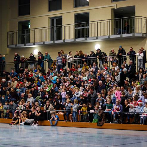 Große Gruppe von Menschen, hauptsächlich Kinder und Eltern, sitzt in einer Sporthalle und schaut auf eine Veranstaltung.