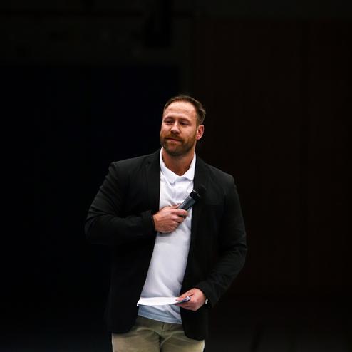 Mann mit Mikrofon in schwarzem Blazer und weißem Polohemd, spricht auf einem Sportplatz mit grünen Linien im Hintergrund.