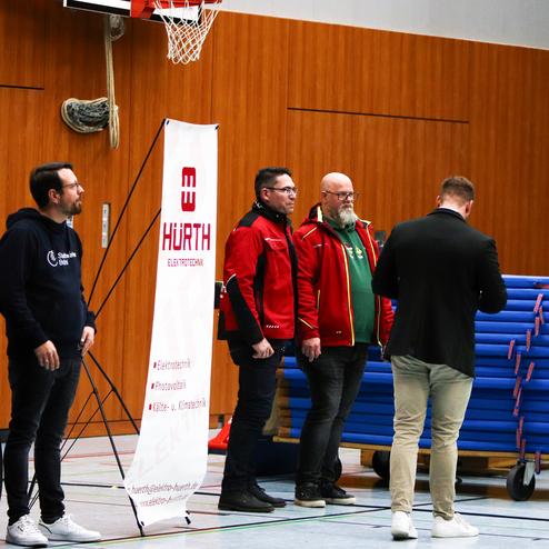 Fünf Personen stehen in einer Turnhalle neben einem Banner mit dem Schriftzug „Hürth Elektrotechnik“ und gestapelten Tischtennisplatten.