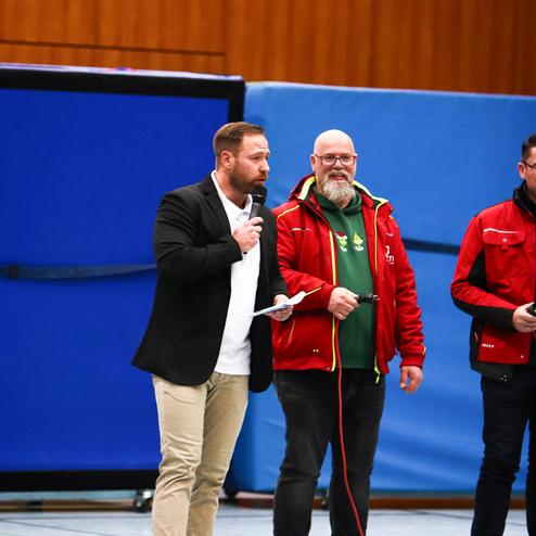 Vier Männer stehen in einer Sporthalle, einer spricht ins Mikrofon, zwei halten Kabel und sie tragen verschiedene Oberteile.