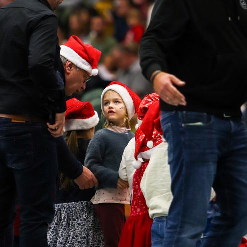 Kinder in Weihnachtsmützen stehen auf einem Event, während ein Erwachsener mit ihnen spricht und sie aufmerksam zuhören.