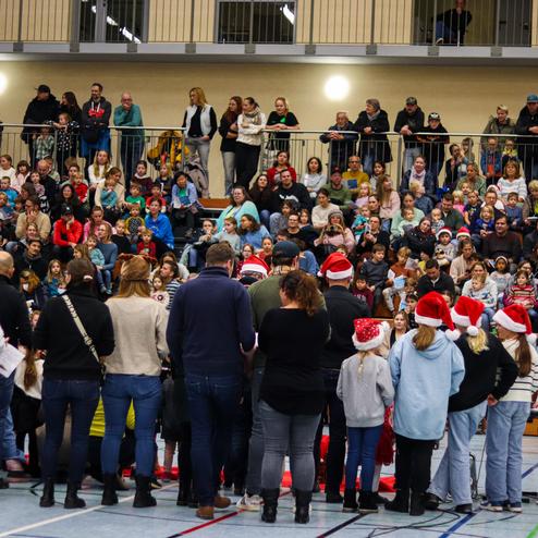 Weihnachtsaufführung mit wachsendem Publikum in einer Halle; Sänger stehen im Vordergrund, viele Kinder und Erwachsene im Hintergrund.