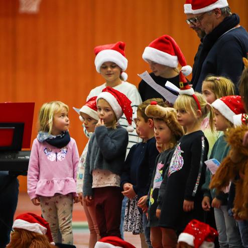Kinder in Weihnachtsmützen singen vor einer Klavierbegleitung in einer Sporthalle. Im Hintergrund Erwachsene lauschen.