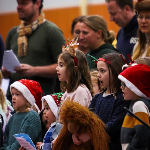 Kinderchor mit Weihnachtsmützen singt in einer Halle, einige halten Plakate und Stofftiere.