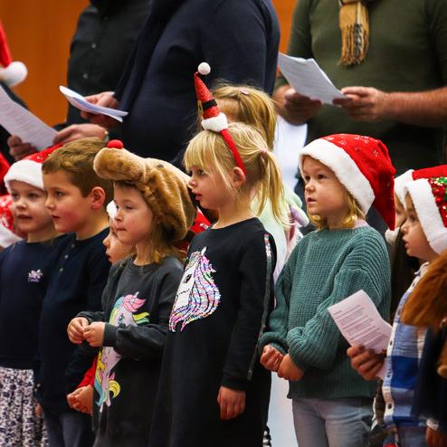 Gruppe von Kindern mit Weihnachtsmützen singt, einige halten Texte in der Hand, vor ihnen stehen Erwachsene.