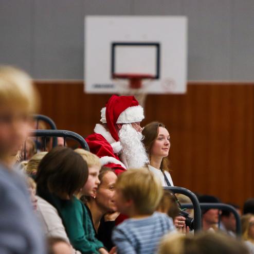 Weihnachtsmann in einem Sporthallen-Bereich, umgeben von Kindern und Erwachsenen, alle erwarten eine Veranstaltung.