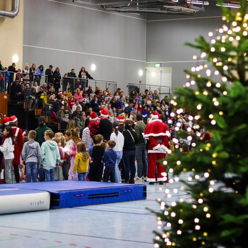 Weihnachtsfeier mit Kindern und Erwachsenen in einer Sporthalle, im Vordergrund ein geschmückter Tannenbaum.