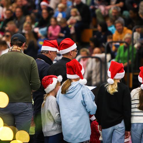Gruppe von Menschen mit Weihnachtsmützen steht auf einer Bühne während einer festlichen Veranstaltung, Publikum im Hintergrund.