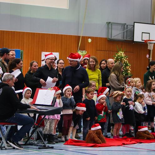 Weihnachtliche Aufführung in einer Halle mit Kindern, Erwachsenen und Weihnachtsmann, um einen Keyboardspieler versammelt.
