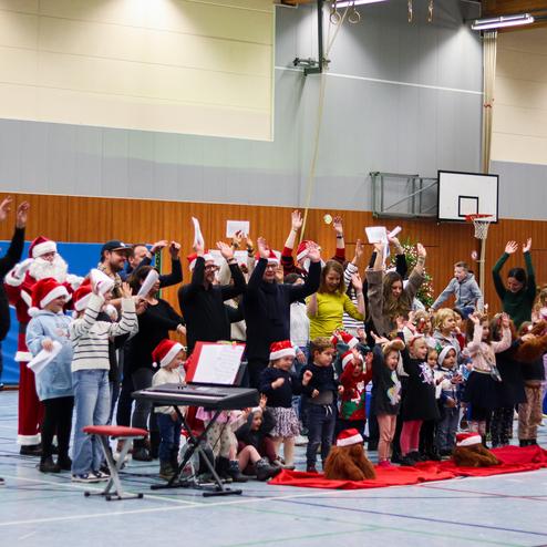 Gruppen von Kindern und Erwachsenen in Weihnachtsmützen singen in einer Sporthalle, umgeben von festlicher Dekoration.