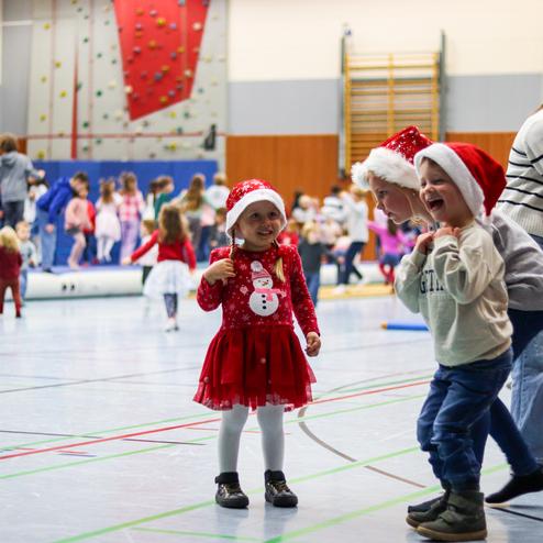 Kinder mit Weihnachtsmützen tanzen in einer Sporthalle, umgeben von anderen spielenden Kindern.