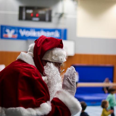 Weihnachtsmann mit rotem Gewand und weißem Bart steht in einer Sporthalle und winkt Kindern zu.