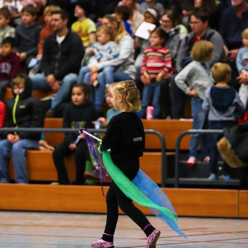 Ein Mädchen mit Zopf trägt ein bunten Tuch und läuft entlang der Tribüne in einer Sporthalle voller Zuschauer.