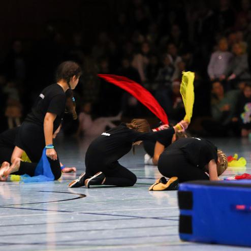 Gruppe junger Sportlerinnen in schwarzen Trikots führt eine Bodenturn-Übung mit bunten Tüchern auf einer Turnfläche aus.