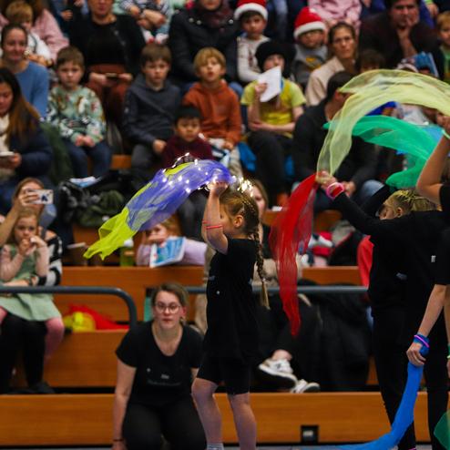 Mädchen tanzt mit bunten Tüchern auf einer Bühne vor einem Publikum in einer Sporthalle.