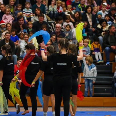 Gruppe von Tänzerinnen mit bunten Tüchern, umgeben von einer Zuschaueransammlung in einer Sporthalle.