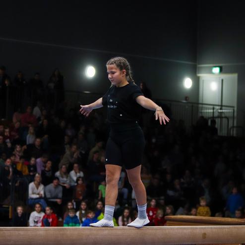 Junge Sportlerin balanciert auf einem Schwebebalken, während eine große Menge im Hintergrund zuschaut.