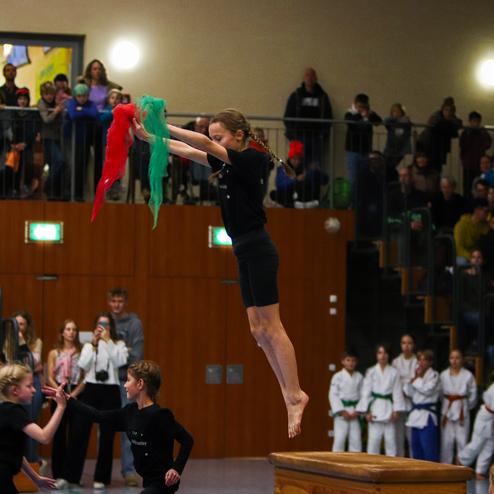 Junge Springer in schwarzer Kleidung, die bunte Tücher hält, springt über eine Turnbank vor einem Publikum.
