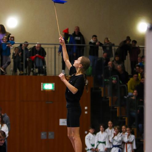 Ein Mädchen balanciert auf einem Kasten und dreht einen blauen Teller auf einem Stock, umgeben von Zuschauern.