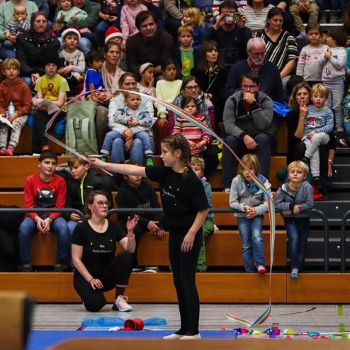 Ein Mädchen führt mit einem bunten Band eine Choreografie vor einer zuschauenden Menschenmenge in einer Sporthalle auf.