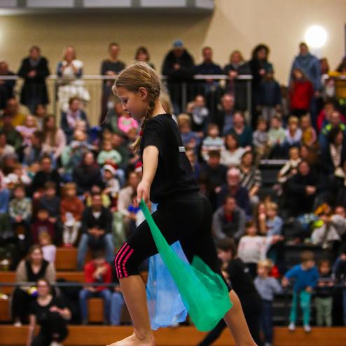 Ein Mädchen mit Zöpfen balanciert auf einem Schwebebalken und hält bunte Tücher in der Hand, umgeben von Zuschauern.