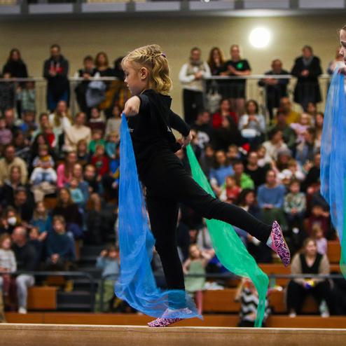 Zwei Mädchen in schwarzer Kleidung führen mit bunten Tüchern eine Choreografie vor einem Publikum in einer Sporthalle auf.