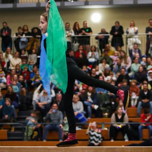 Mädchen führt eine Tanzroutine auf einer Bühne mit bunten Tüchern vor einer Zuschauergruppe in einer Sporthalle auf.
