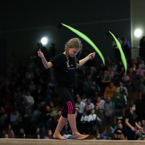 Mädchen mit Zöpfen und bunten Bändern jongliert auf einem Schwebebalken vor einem Publikum in einem Sporthalle.