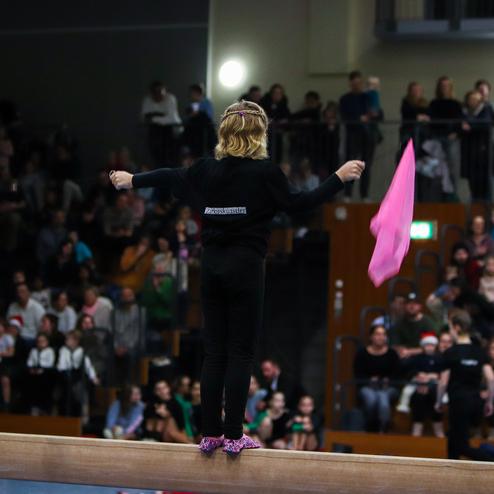 Mädchen balanciert auf einem Balken und schwenkt pinke Tücher vor einem Publikum in einem Festsaal.