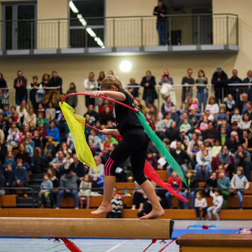 Mädchen balanciert auf einem Schwebebalken und schwingt farbige Tücher vor einem Publikum in einer Turnhalle.