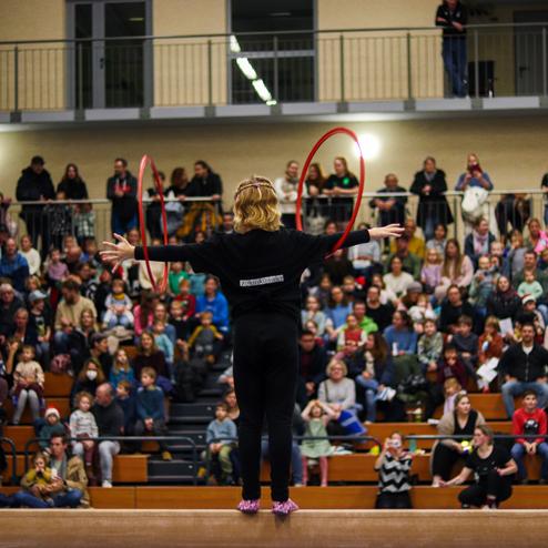 Junge Artistin mit Hula-Hoops und bunten Tüchern auf der Bühne vor einem großen Publikum in einer Turnhalle.
