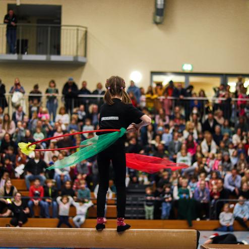 Mädchen auf einem Sprungbrett wirkt mit bunten Tüchern vor einem Publikum in einer Sporthalle.