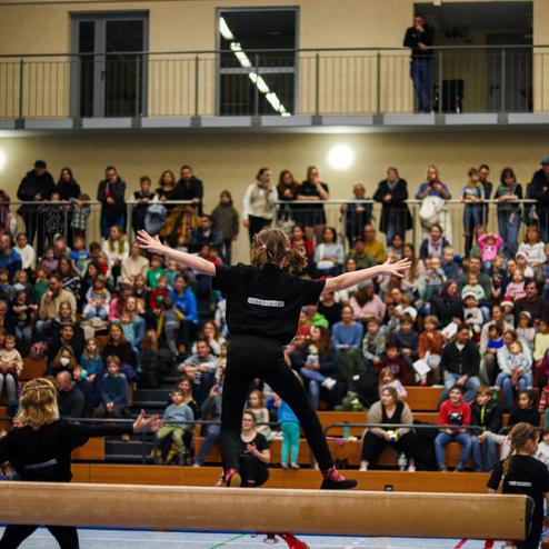 Ein Mädchen turnt mit ausgebreiteten Armen auf einem Schwebebalken vor einer großen Zuschauergruppe in einer Sporthalle.