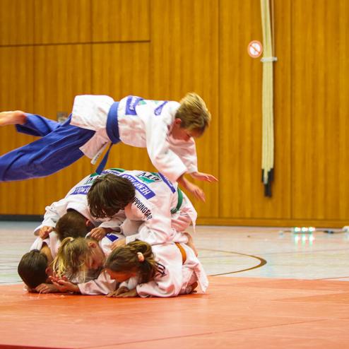 Junge mit blauer Judo-Gi springt über eine Gruppe von anderen Kindern in weißen Judokas auf einer Judo-Matte.