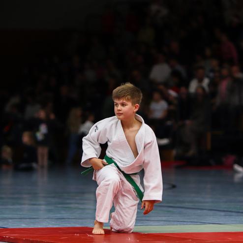 Junge in Judokleidung mit grünem Gürtel kniet auf einer rot-grünen Mattenfläche in einer Sporthalle.