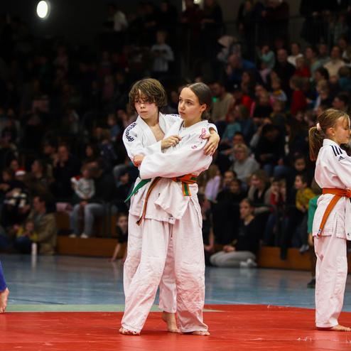 Zwei Jugendliche im Judoanzug stehen auf einer roten Matte, umgeben von Zuschauern in einem Wettkampf-Ambiente.