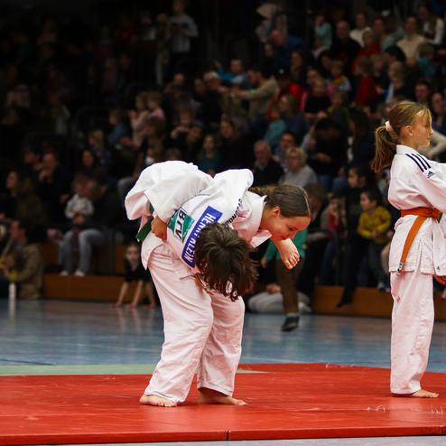 Vier Kinder in Judoanzügen trainieren auf einer roten Matte, während eine Menschenmenge zuschaut.