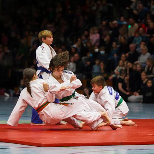 Junge Kämpfer im Judoanzug führen einen Wurf auf einer roten Matte vor einer zuschauenden Menge aus.