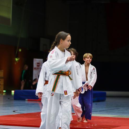 Vier Kinder in Judoanzügen stehen auf einer roten Matte in einer Sporthalle während des Trainings.