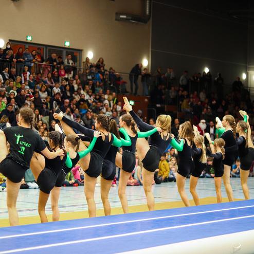 Gruppenvorführung von Kunstturnerinnen mit erhobenen Beinen auf einer Turnmatte, Publikum im Hintergrund.