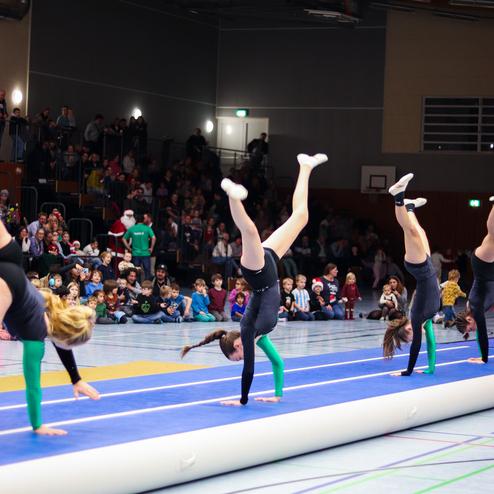 Vier Turnerinnen führen Handstände auf einer blauen Turnmatte vor einem Publikum in einer Sporthalle aus.