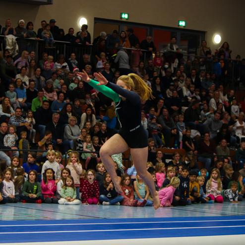 Gymnastin springt auf einer blauen Turnmatte vor einer jubelnden Zuschauermenge, überwiegend Kinder und Familien.