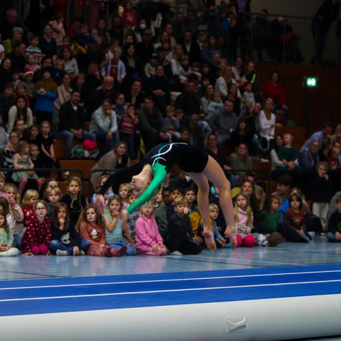 Junge Sportlerin in einem schwarzen Trikot springt über eine weiche Aufsprungfläche vor einem Publikum aus Kindern und Erwachsenen.