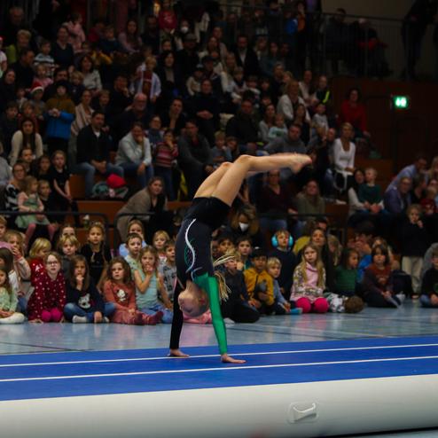 Junge Sportlerin führt einen Handstand auf einer blauen Turnmatte vor einer zuschauenden Menge aus.