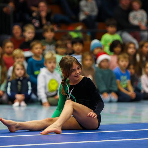 Ein Mädchen in einem schwarzen Gymnastikanzug führt eine Übung auf einer blauen Matte vor aufmerksamen Zuschauern aus.