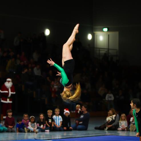 Turnerin führt einen Salto in der Luft, während Zuschauer im Hintergrund auf einer Gymnastikmatte gespannt zusehen.