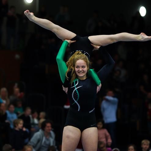 Zwei Turnerinnen in Bodensynchronisation; eine hebt die andere in der Luft, umgeben von Zuschauern in der Arena.