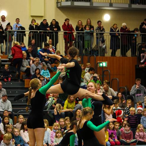 Fünf Turnerinnen führen eine pumpende Übung in einem vollen Sporthallenvorführung vor, Zuschauer beobachten aufmerksam.