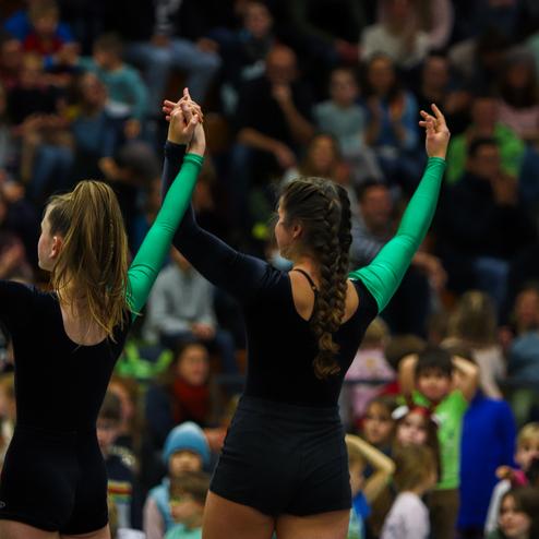 Drei Mädchen in Sportanzügen mit grünen Ärmeln zeigen ihre Hände Richtung Publikum bei einer Veranstaltung.