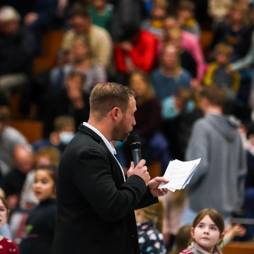 Mann mit Mikrofon und Unterlagen spricht vor Publikum, Kinder bewegen sich im Vordergrund.